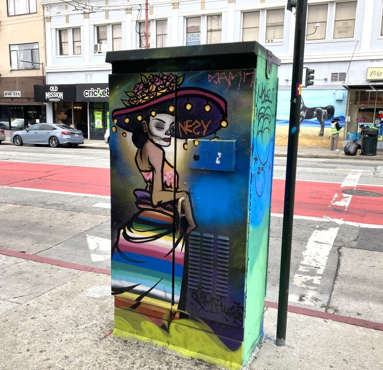 Colorful street art on a utility box depicts a seated skeleton figure in a large hat with flowers, set against a city street background with shops and a mural.