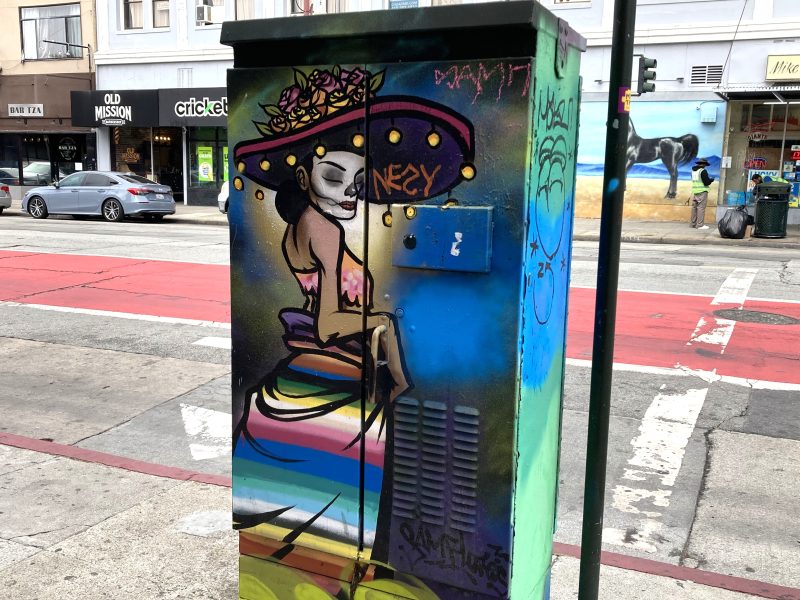 Colorful street art on a utility box depicts a seated skeleton figure in a large hat with flowers, set against a city street background with shops and a mural.