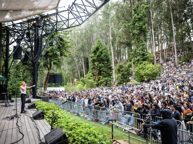A performer on an outdoor stage addresses a large audience seated and standing among trees in the Stern Grove amphitheater setting.