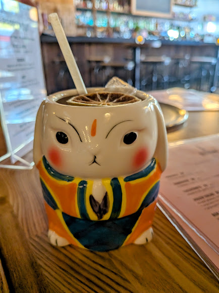 A cocktail served in a ceramic cup shaped like a rabbit, with a straw and a dried citrus slice on top, placed on a wooden table in a bar setting.