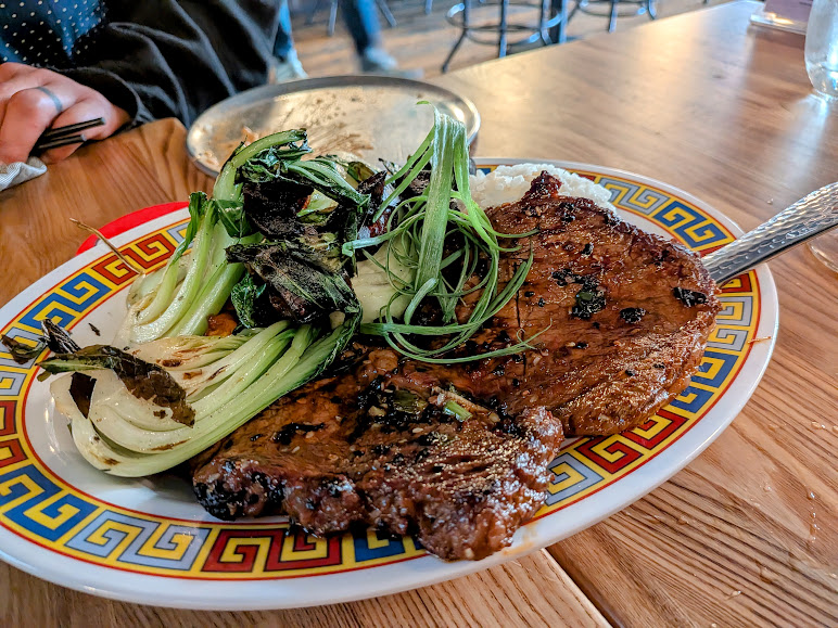 A plate of grilled steak with greens and rice is served on a decorative dish on a wooden table, with a person sitting nearby.