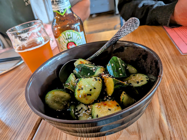 A bowl of sliced cucumbers topped with sesame seeds sits on a wooden table next to a glass of beer and a bottle.
