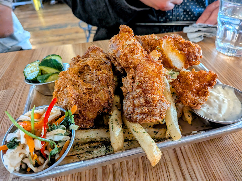 A tray with battered fried fish on top of seasoned fries, served with tartar sauce, pickles, and coleslaw on a wooden table.