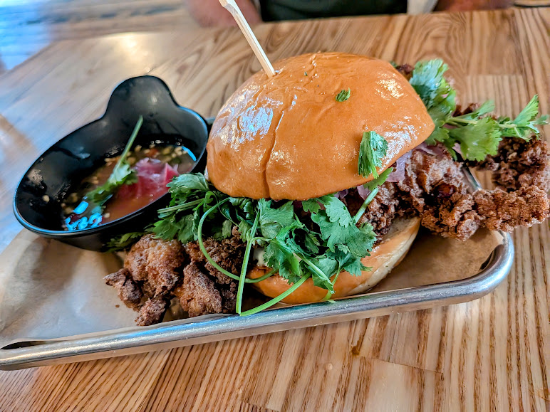 A fried soft-shell crab sandwich with fresh cilantro on a brioche bun, served with a side of dipping sauce on a metal tray.