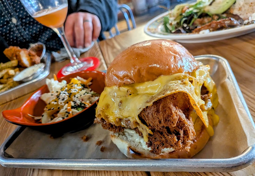 A fried chicken sandwich with melted cheese on a bun, served with a side of slaw and fries on a metal tray. A person and a glass of beverage are in the background.