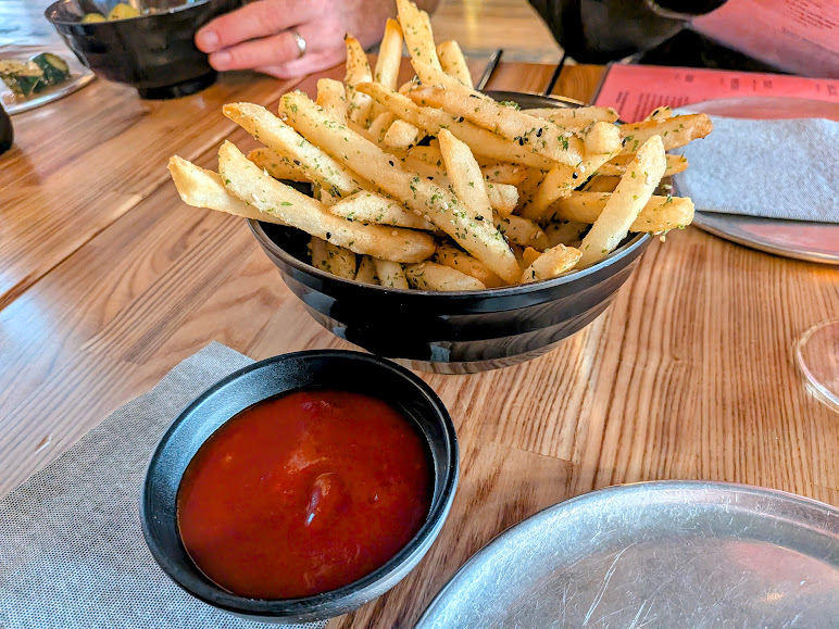A black bowl filled with seasoned French fries is on a wooden table, next to a small black dish of ketchup.