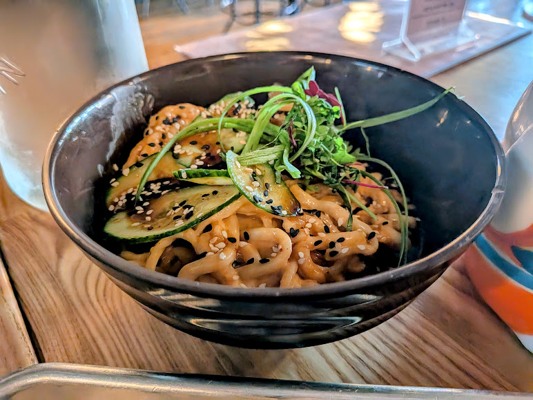 A black bowl filled with noodles topped with sliced cucumber, greens, and a sprinkling of black and white sesame seeds, placed on a wooden table.