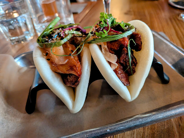 Two steamed bao buns filled with sliced meat, vegetables, and garnished with herbs, served on a metal tray with a glass of water in the background.