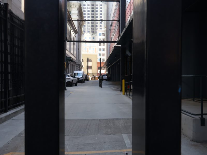 A person walks down a city alleyway lined with parked cars and tall buildings, viewed through a black metal gate.
