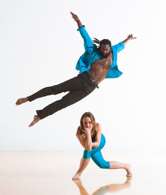A man in a blue shirt and black pants leaps in the air above a woman in blue athletic wear who is kneeling on the floor, set against a plain white background.