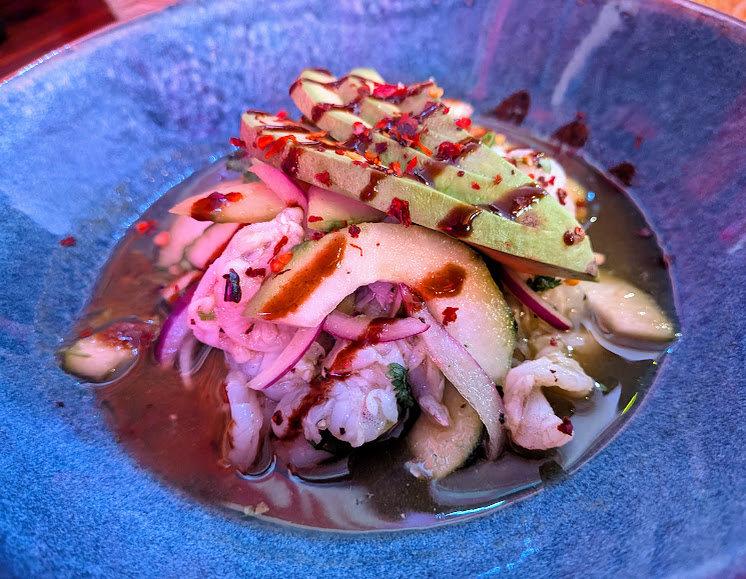 A blue bowl filled with ceviche topped with thinly sliced avocado, cucumber, red onion, and red chili flakes in a citrus-based sauce.