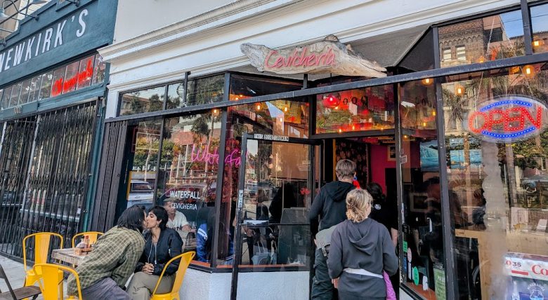 People are seated at yellow tables outside and standing at the entrance of a restaurant called "Calçheria," with an "OPEN" sign in the window.