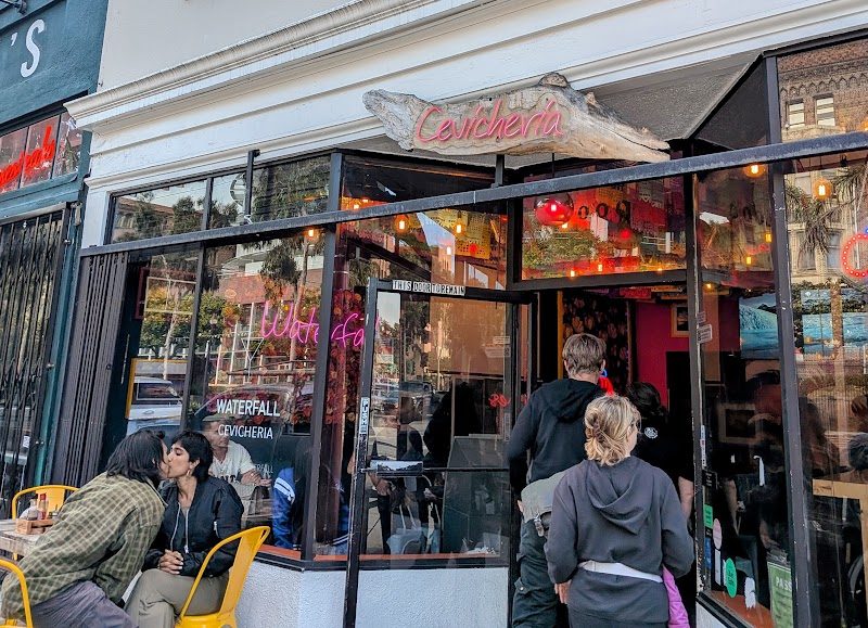 People are seated at yellow tables outside and standing at the entrance of a restaurant called "Calçheria," with an "OPEN" sign in the window.