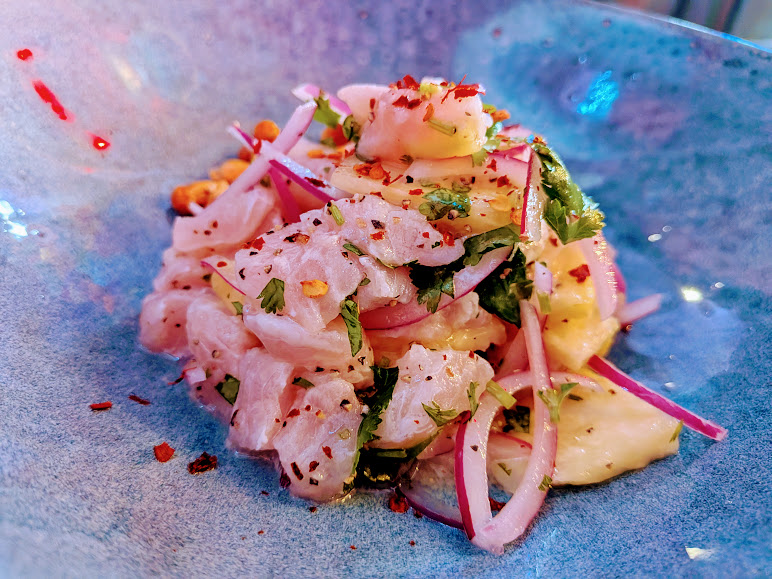 A close-up of a ceviche dish with raw fish, sliced red onions, cilantro, and red chili flakes served on a blue plate.