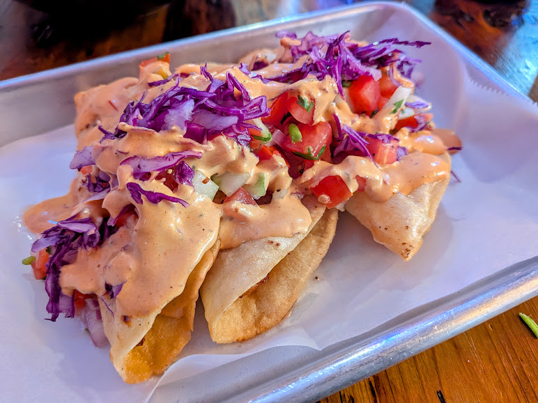 Three crispy tacos filled with fish, topped with diced tomatoes, onions, shredded purple cabbage, and drizzled with a creamy orange sauce, served on a tray.