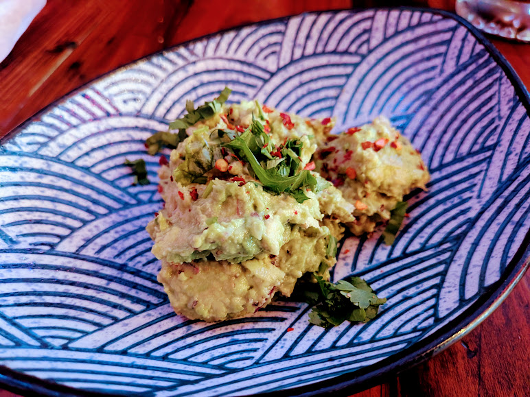 A patterned blue and white plate with three scoops of guacamole, garnished with chopped cilantro and red pepper flakes, on a wooden table.