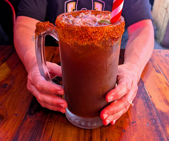 A person holding a large glass mug filled with a dark drink, ice, and a red-and-white striped straw; the rim is coated with chili powder.