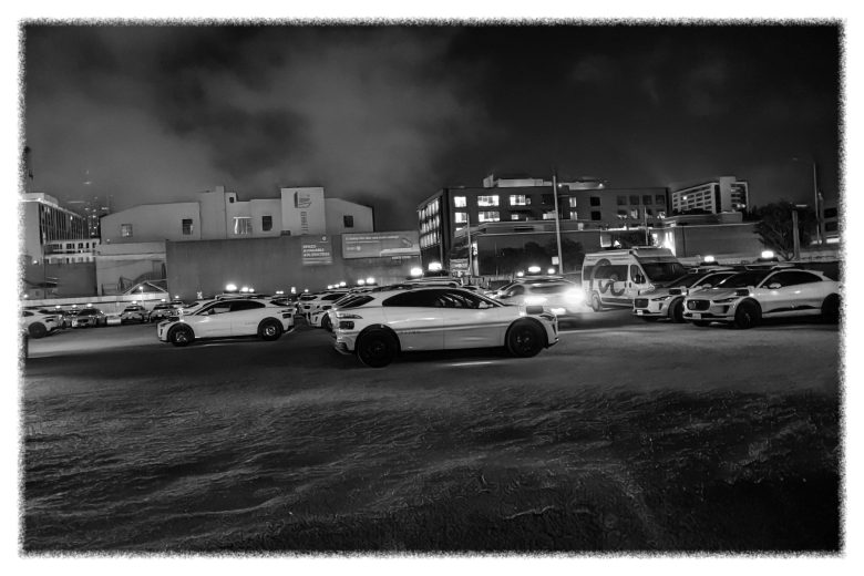 A parking lot filled with cars at night, surrounded by buildings under a cloudy sky. The scene is in black and white.