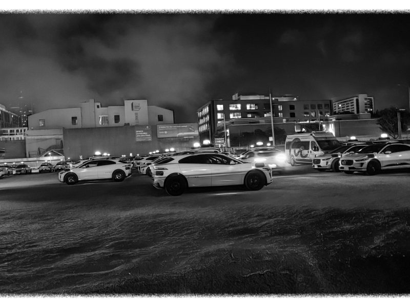 A parking lot filled with cars at night, surrounded by buildings under a cloudy sky. The scene is in black and white.