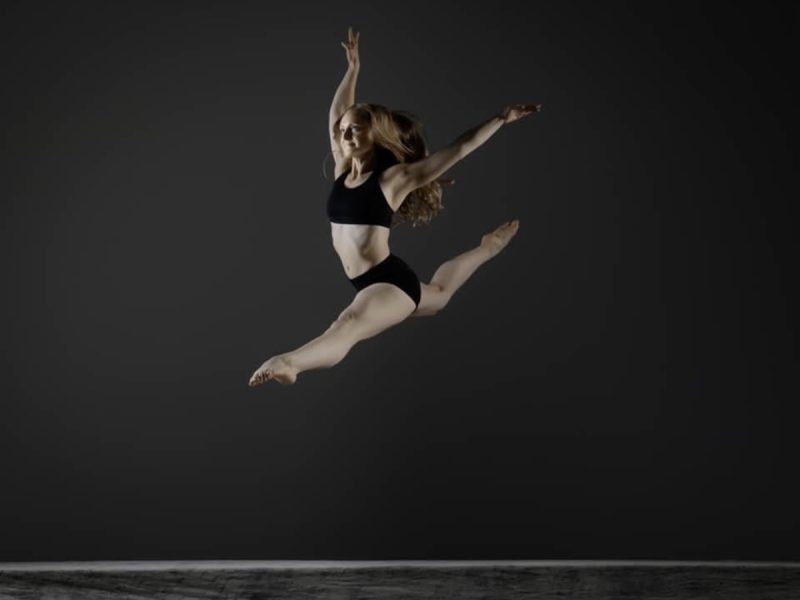 A dancer in a black outfit performs a split leap in mid-air against a dark background.