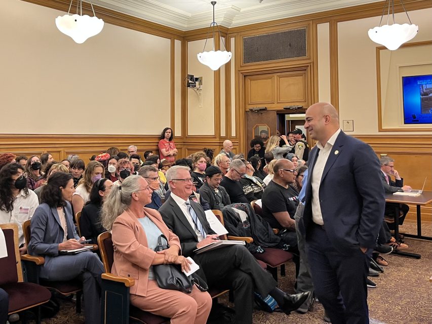 A man in a suit stands and smiles at the front of a crowded room where people are seated and appear to be attending a formal meeting or hearing.