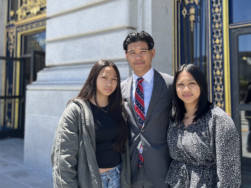 Three people stand together outside a building with ornate gold and black doors, one wearing a suit and tie, and the others in casual attire.