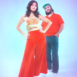 A woman in a strapless gold top and red pants stands in front of a man wearing a red shirt, green pants, and a green cap, both posing against a blue background.