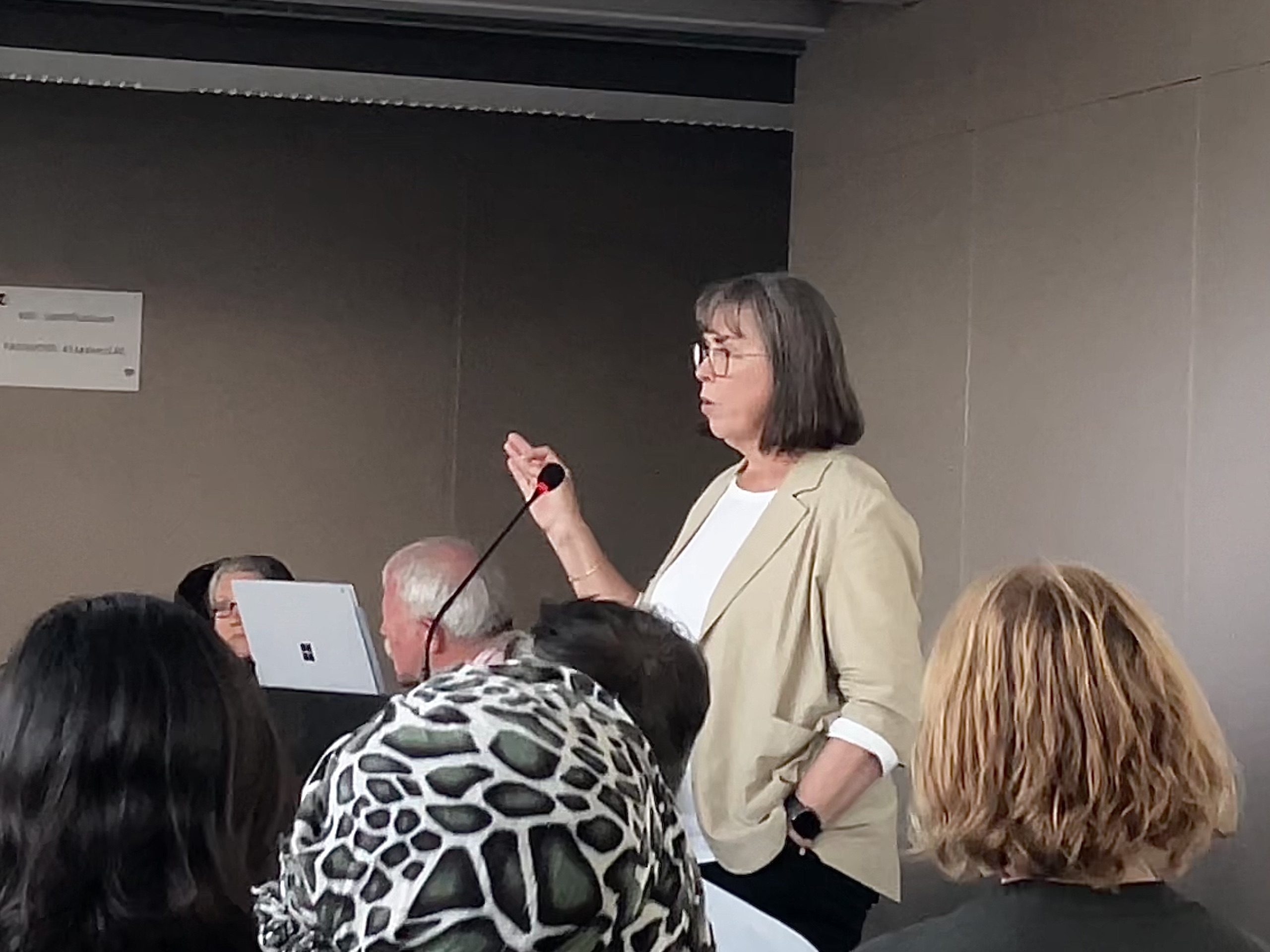 A woman stands and speaks into a microphone in a conference room, addressing a seated audience.