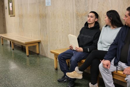 Three people sit on a bench in a hallway; the person in the middle, possibly facing a teen prison sentence, holds a folder and looks to the left, while the others look straight ahead.