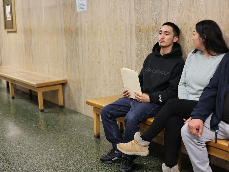 Three people sit on a bench in a hallway; the person in the middle, possibly facing a teen prison sentence, holds a folder and looks to the left, while the others look straight ahead.