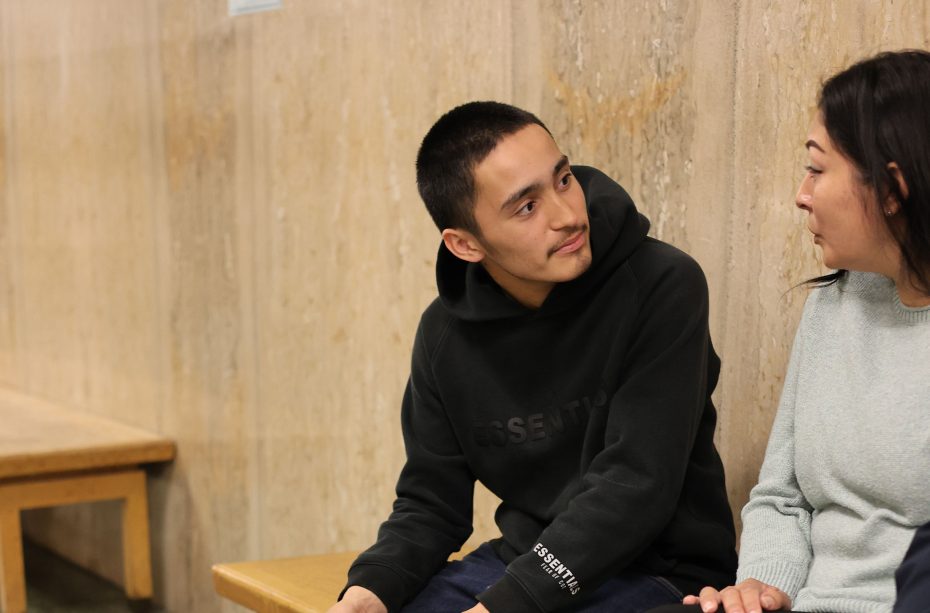 A young man in a black hoodie sits on a bench, sharing with a woman in a light sweater about his teen prison sentence as they talk against a beige wall.