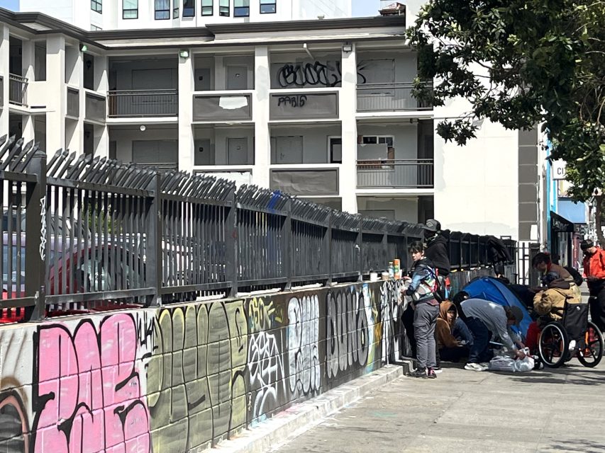 A group of people gather on a sidewalk beside a graffiti-covered fence, near an apartment building. One person is in a wheelchair.