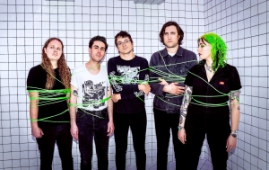 Five people stand against a tiled wall, partially wrapped with green string, looking toward the camera.