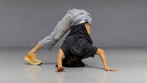 A person in jeans and a black shirt performs a breakdancing move, balancing on their head and hands with legs bent and feet on the floor.