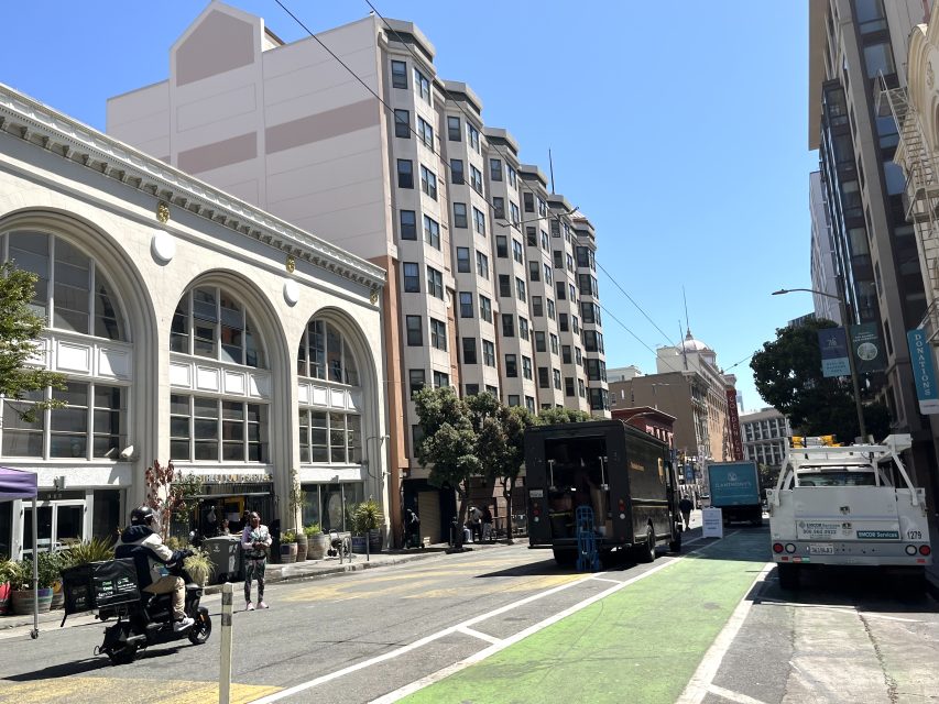 City street with a bike lane, delivery trucks, pedestrians, and a person riding a motorized scooter in front of an arched building on a sunny day.