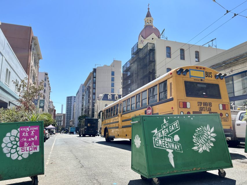 A yellow school bus and green dumpsters are on a city street lined with buildings under a clear blue sky.