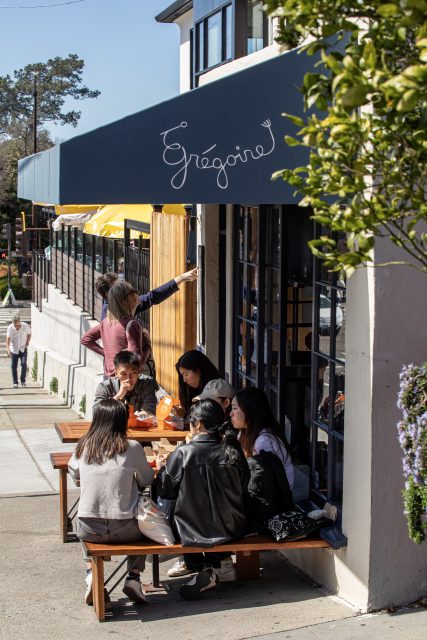 A group of people sits at a wooden table outside a café named Grégoire on a sunny day.