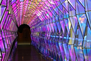 A tunnel with geometric glass panels illuminated by purple, pink, and blue lights, creating a vibrant, reflective passageway.