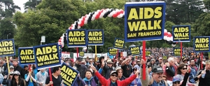 A large crowd participates in the AIDS Walk San Francisco, holding matching signs and walking under a red, white, and black balloon arch in a park setting.