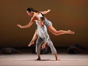 A woman in a white outfit balances on the back of a man in gray during a contemporary dance performance on stage with a neutral background.