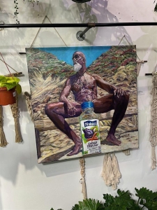 A painting of a seated man outdoors with a large Naked juice bottle digitally added over his lower body, displayed on a wall among hanging plants and macramé decor.