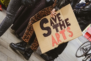 Person holding a cardboard sign reading "Save the Arts" during a public demonstration; legs and shoes of participants visible on pavement.