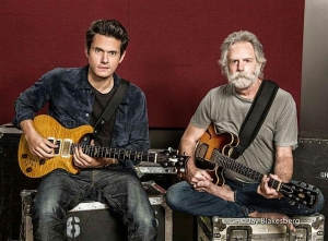 Two men sit side by side holding electric guitars in front of a red and gray wall, with musical equipment cases around them.