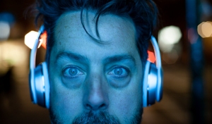A man with blue-lit headphones looks directly at the camera at night, with a serious expression and bright city lights blurred in the background.