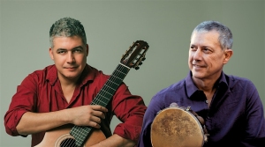 Two men are shown; one holds a classical guitar and wears a red shirt, while the other holds a tambourine and wears a purple shirt. Both are seated against a plain background.