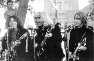 Three musicians perform outdoors with guitars and microphones in front of a crowd.