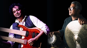 Two men are shown against a black background; one plays a double-neck electric guitar, while the other holds an Indian percussion instrument, possibly a tabla or mridangam.