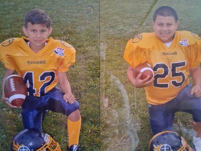 Two boys in yellow football uniforms kneel on grass, each holding a football. Their helmets are on the ground in front of them. The boy on the left has jersey number 42, the boy on the right number 25.