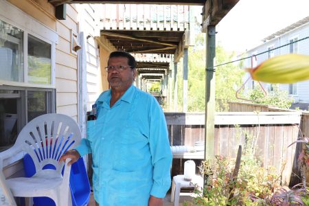 A man in a blue shirt stands on a porch holding a white plastic chair, with houses and plants visible in the background.