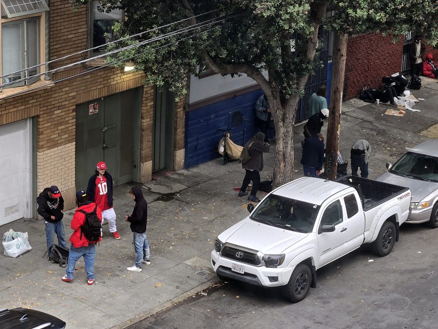 Several people stand and sit on a city sidewalk near a parked white pickup truck, with some gathered in groups and others near a building and trees.